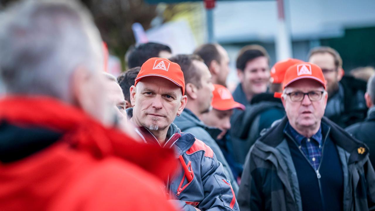 Ein Mann mit Arbeitsjacke und roter Kappe blickt in das Gesicht eines Mannes am Bildrand.