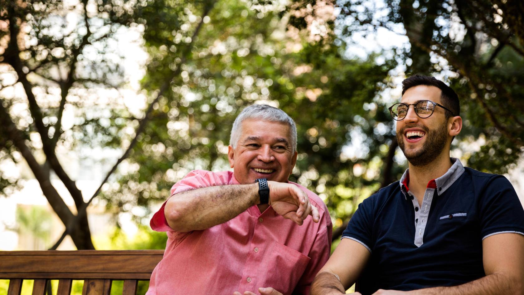 Ein junger und ein älterer Mann sitzen lachend nebeneinander auf einer Bank.